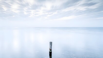 Minimalist long exposure seascape featuring a single wooden post in calm blue water under a soft overcast sky with misty horizon, copy space area