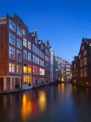 Naklejka premium Beautiful Amsterdam canal at twilight with historic dutch architecture. Picturesque evening view of Amsterdam historical district at dusk, featuring glowing windows and calm water reflections.