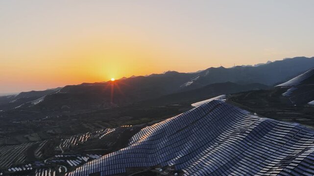 Aerial video of a mountain solar farm at sunset, with golden light illuminating rows of photovoltaic panels across rolling hills.