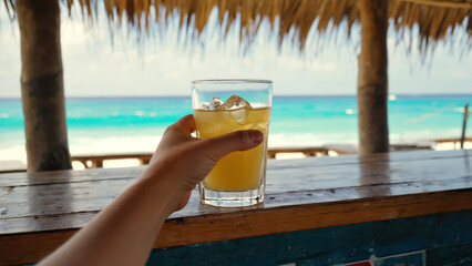 A hand is holding a cup filled with pineapple juice and ice cubes. A tropical beach bar serves as the backdrop, with palm shadows and ocean waves