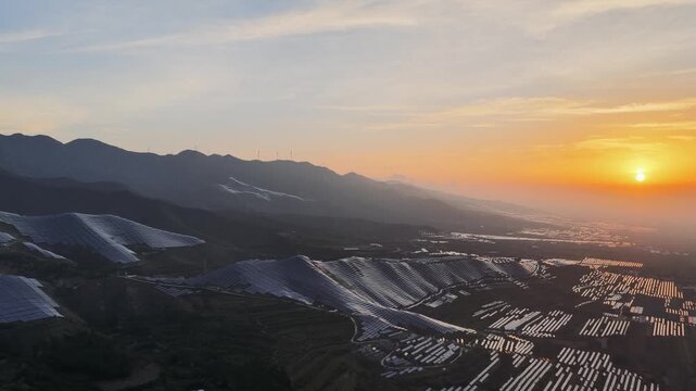 Aerial footage of a mountain solar farm at sunrise, with golden light illuminating rows of photovoltaic panels across rolling hills.