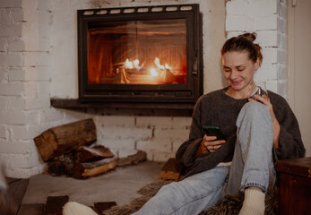 Young woman by the fireplace in a cozy house with a mobile phone in her hands
