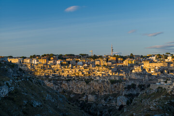 Fototapeta premium Soft golden light bathes the layered stone buildings of Matera, Italy, highlighting the dramatic cliffside setting beneath a clear blue sky with scattered clouds.