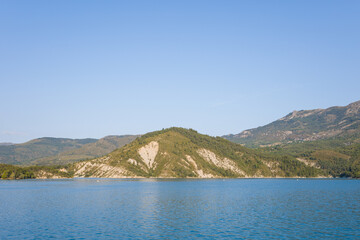 Obraz premium Serene view of a forested hill rising above the tranquil waters of Lac de Castillon, with rugged slopes and distant mountains under a clear blue sky.