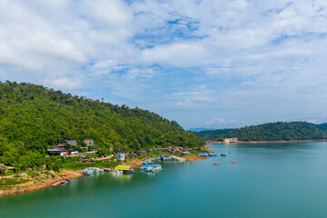 Fototapeta premium Cluster of colorful boats docked beside a lush green hillside on the banks of Nam Ngum Reservoir in Laos. Calm turquoise water and scattered clouds create a tranquil lakeside atmosphere surrounded by