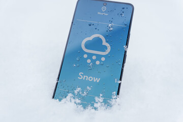 Smartphone lying in snow with snow forecast on weather app screen, winter outdoors concept