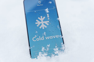 Smartphone lying in snow with cold wave alert on weather app screen, winter outdoors concept