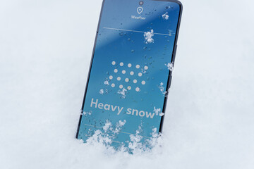 Smartphone standing in snow with heavy snow forecast on weather app screen, winter storm concept
