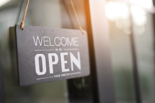 Black open sign with welcome text hanging on glass door of modern cafe, showing business opening hours and customer invitation. Vintage color tone style.
