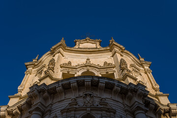 Fototapeta premium Upward view of an ornate baroque church facade in Lecce, Italy, highlighted by warm sunlight against a deep blue sky. The intricate stonework and sculpted details showcase the architectural richness