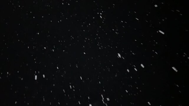 thick snow falls at night in the light of a streetlamp, snowflakes swirl in the wind and rush away against a black background.