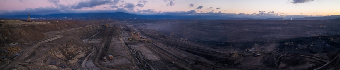High resolution panoramic view of an open pit coal mine with mining machines, layered terrain. Industrial landscape showing large scale extraction, energy production, and environmental impact.