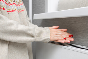  A girl in a knitted sweater warms her hands near a radiator. A girl warms herself near a heating radiator. person warming up. heating season. cold season. heating radiator