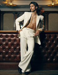 Sophisticated gentleman in flowing white suit poses confidently against rich leather interior with dramatic golden lighting