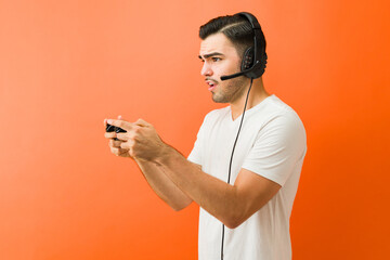 Profile view of a man concentrating on playing video game with controller and headset against an orange background