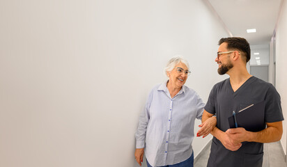 Male doctor walking arm in arm with senior woman in hospital lobby during discussion about health concerns and plan
