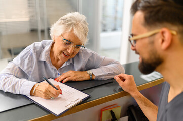Male receptionist looking at senior patient filling form of medical document at reception desk in hospital