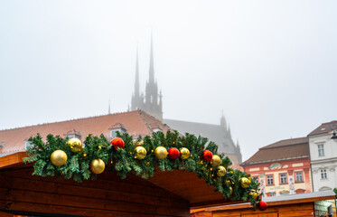 Decorated roof of a christmas market stall with fir branches and red and gold ornaments, brno city atmosphere with foggy silhouette of peter and paul cathedral in background