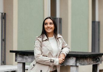 Smiling woman with long dark hair wearing a stylish beige trench coat, leaning on a wooden table, set against a modern urban backdrop, showcasing contemporary fashion and confidence