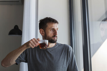 Adult man in casual t shirt standing by window and drinking tea from glass cup at home, calm morning mood, real people lifestyle, thoughtful expression, natural light, authentic everyday moment