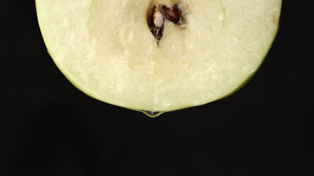 Dark background close-up of juicy apple slice with core and seeds as droplet forms and drips in fresh fruit nutrition context