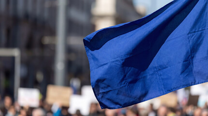 Close-up of a blank blue flag waving in the wind with a blurred crowd of protesters in the background