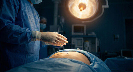 A surgeon in blue scrubs and gloves preparing to operate on a patient on an operating table under a bright surgical lamp.