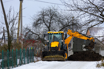 A machine digs and moves dirt on a snowy road in a rural neighborhood while trees stand bare in the cold