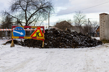 Construction work blocks the road with a dirt pile and traffic signs indicating detours in a snowy landscape during winter