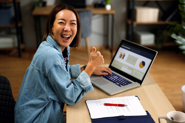 Happy Asian woman showing thumbs up while working on laptop at home