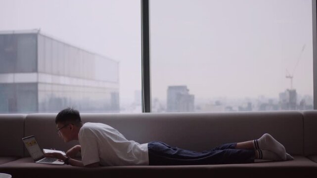 Asian man lying on sofa using laptop near window with city skyline view, remote work lifestyle, relaxed productivity, freelance work from home, modern living, digital nomad and work life balance conce