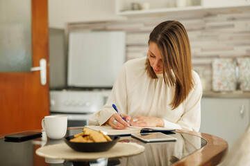 Woman writing notes in planner and using tablet computer while working remotely from home