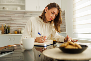 Woman writing notes in planner and using tablet computer while working remotely from home