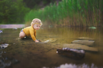 Cute toddler girl with curly hair and flower headband playing in a shallow natural pond. She wears a yellow UV rash guard shirt, crawling and exploring nature on a warm summer day.