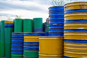 Barrels in various colors are lined up and stacked in a yard with clouds visible in the sky and trees nearby