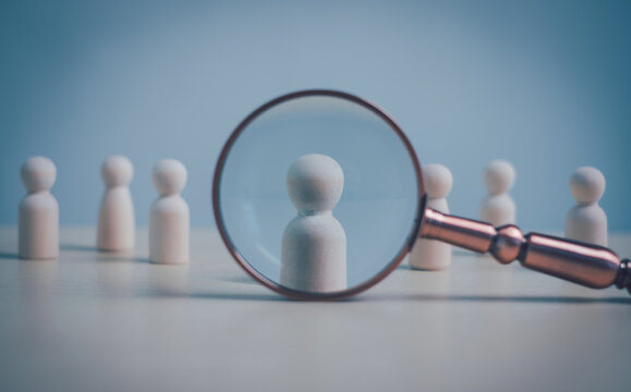 Wooden figures with magnifying glass highlighting one person, talent selection, human resources and leadership concept. Minimal business image for hiring, search and team management.