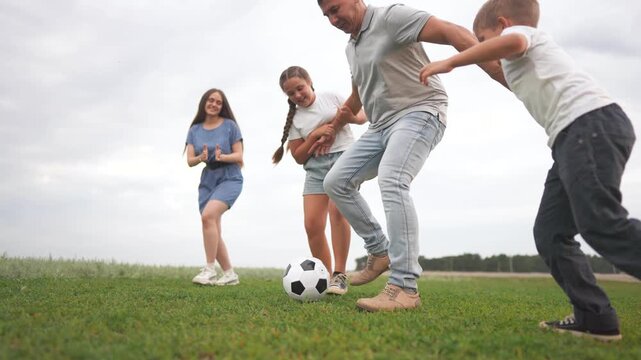 The girl, man, and boy play soccer on a grassy field while the girl runs towards the ball. The man kicks the soccer ball as the boy and girl interact nearby.