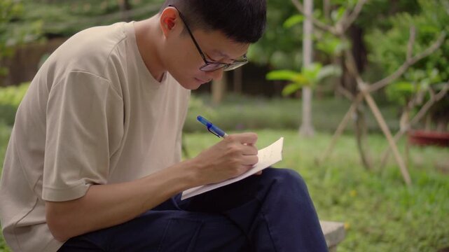Asian man writing notes in notebook outdoors, focused handwriting in park, creative writing and thinking process, calm lifestyle, self reflection and learning concept