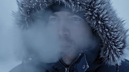 Close-up of a man's face in a fur-lined parka hood exhaling visible breath. Freezing winter weather concept with steam condensing in cold air. Portrait of a male in extreme temperature