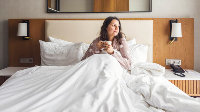 Calm woman sitting in a hotel bed with white bedding, holding a cup of coffee and looking away thoughtfully, cozy morning atmosphere, quiet moment of reflection, lifestyle and emotions concept