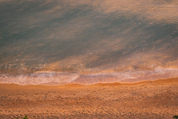 Top-down view of gentle ocean waves washing onto sandy beach with warm sunlight tones, creating a calm natural shoreline texture ideal for background, travel, wellness, and minimal design concepts.