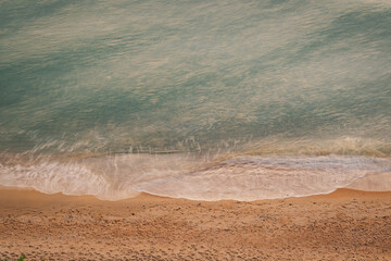 Top-down view of gentle ocean waves washing onto sandy beach with warm sunlight tones, creating a calm natural shoreline texture ideal for background, travel, wellness, and minimal design concepts.