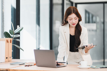 Professional Asian businesswoman using digital tablet and laptop while standing at her desk in a modern office workspace.