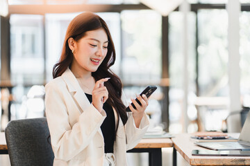 Obraz premium Young Asian businesswoman smiling and using a smartphone while sitting at a desk in a bright modern office with a laptop.