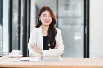 Portrait of a confident young Asian businesswoman smiling with arms crossed while sitting at her modern office desk.