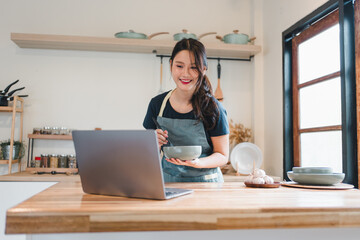 Smiling young Asian woman in an apron holding a bowl while looking at a laptop recipe in a modern kitchen, following an online cooking tutorial.