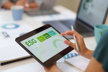 Person's hands holding a tablet and touching an ESG dashboard with data charts while working in a modern office environment.