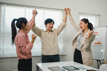 Three happy Asian business people celebrating success and raising hands during an office meeting for teamwork and victory.