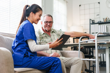 Asian female nurse in blue scrubs showing digital tablet to elderly senior man with walker while sitting on sofa at home.