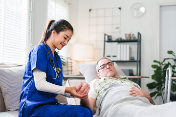 Young Asian nurse in blue scrubs checking the heart rate pulse of a senior male patient lying in bed during a home healthcare visit.